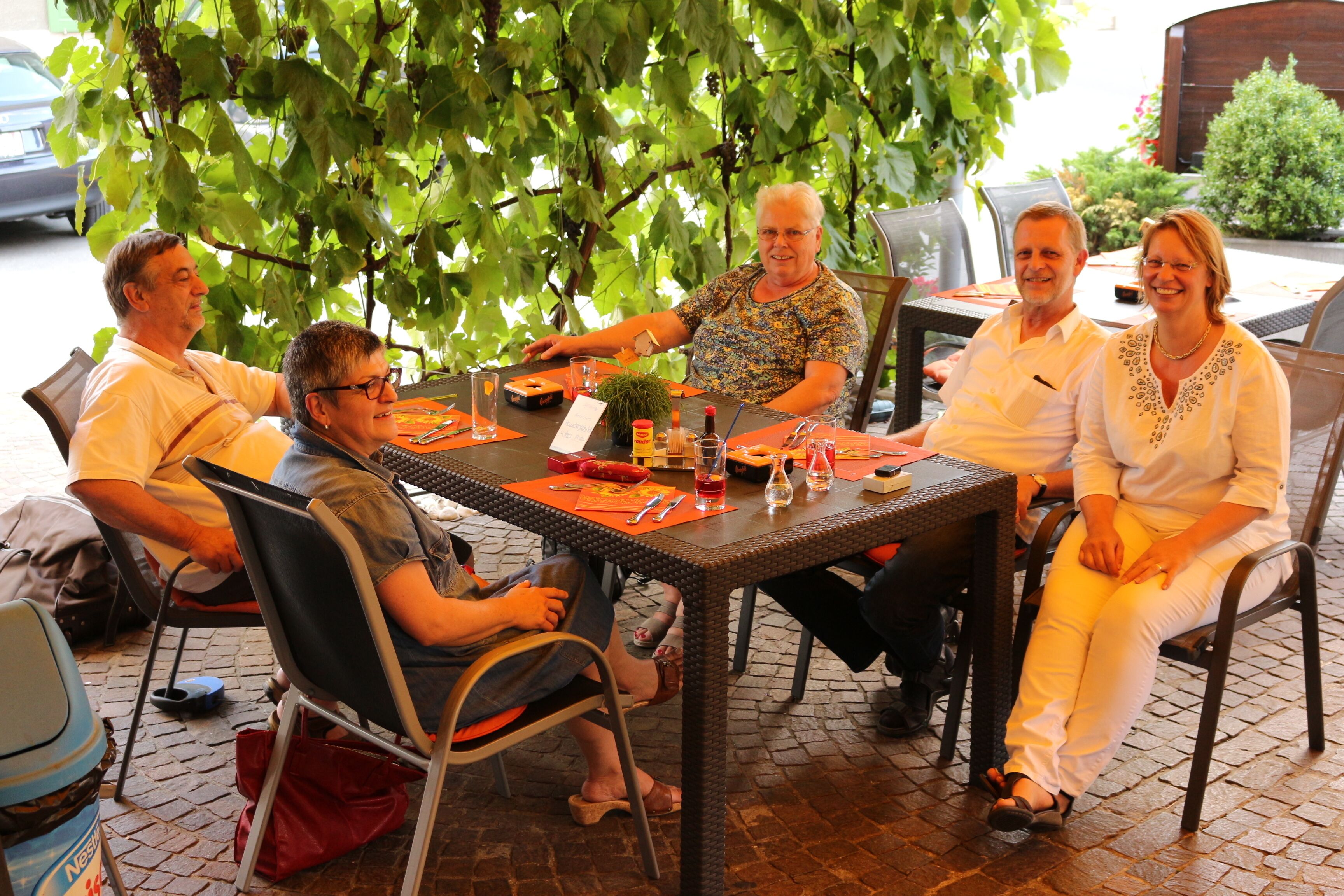 Un groupe de personnes est assis autour d’une table, avec une plante, dans un cadre extérieur où le sol est recouvert de briques.