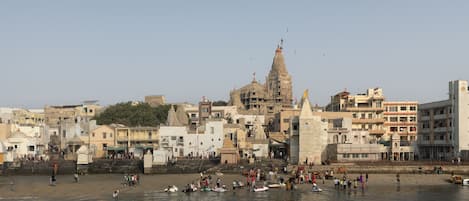 Un paisaje costero con una estructura imponente de estilo templario al fondo, una playa arenosa con gente y barcos, y un cielo despejado.