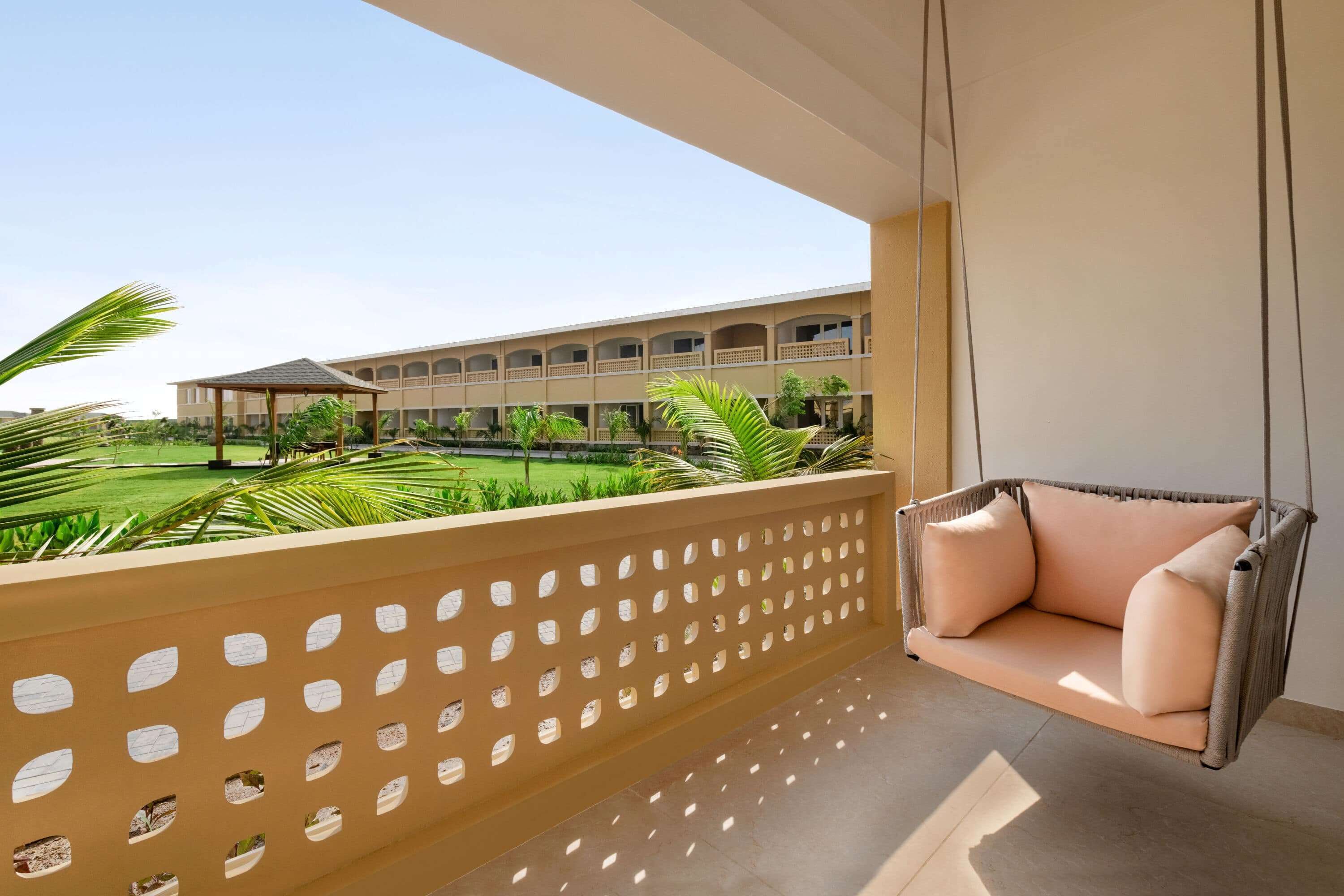 A balcony with a hanging chair, a view of a building with multiple balconies, and a green lawn with palm trees.
