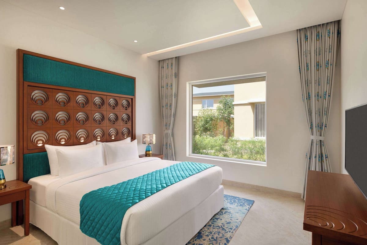 Una habitación de hotel con una cama grande, cabecera de madera, televisor y ventana con cortinas.