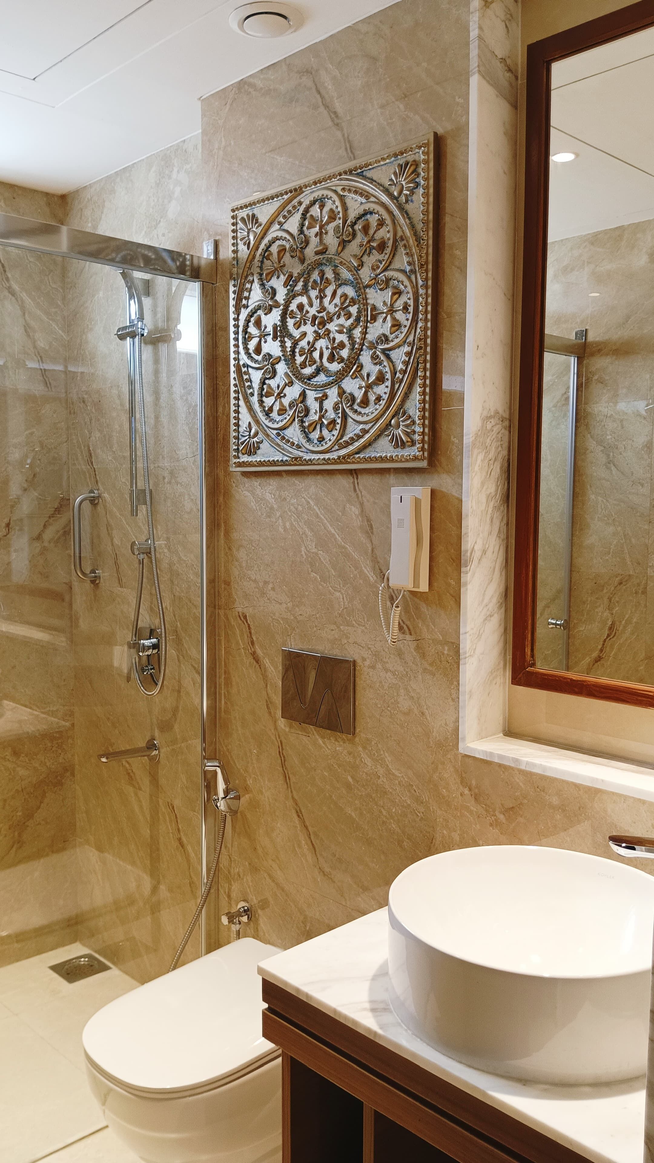 Baño con ducha envuelta en vidrio, inodoro, lavamanos con encimera de mármol y un panel decorativo en la pared.