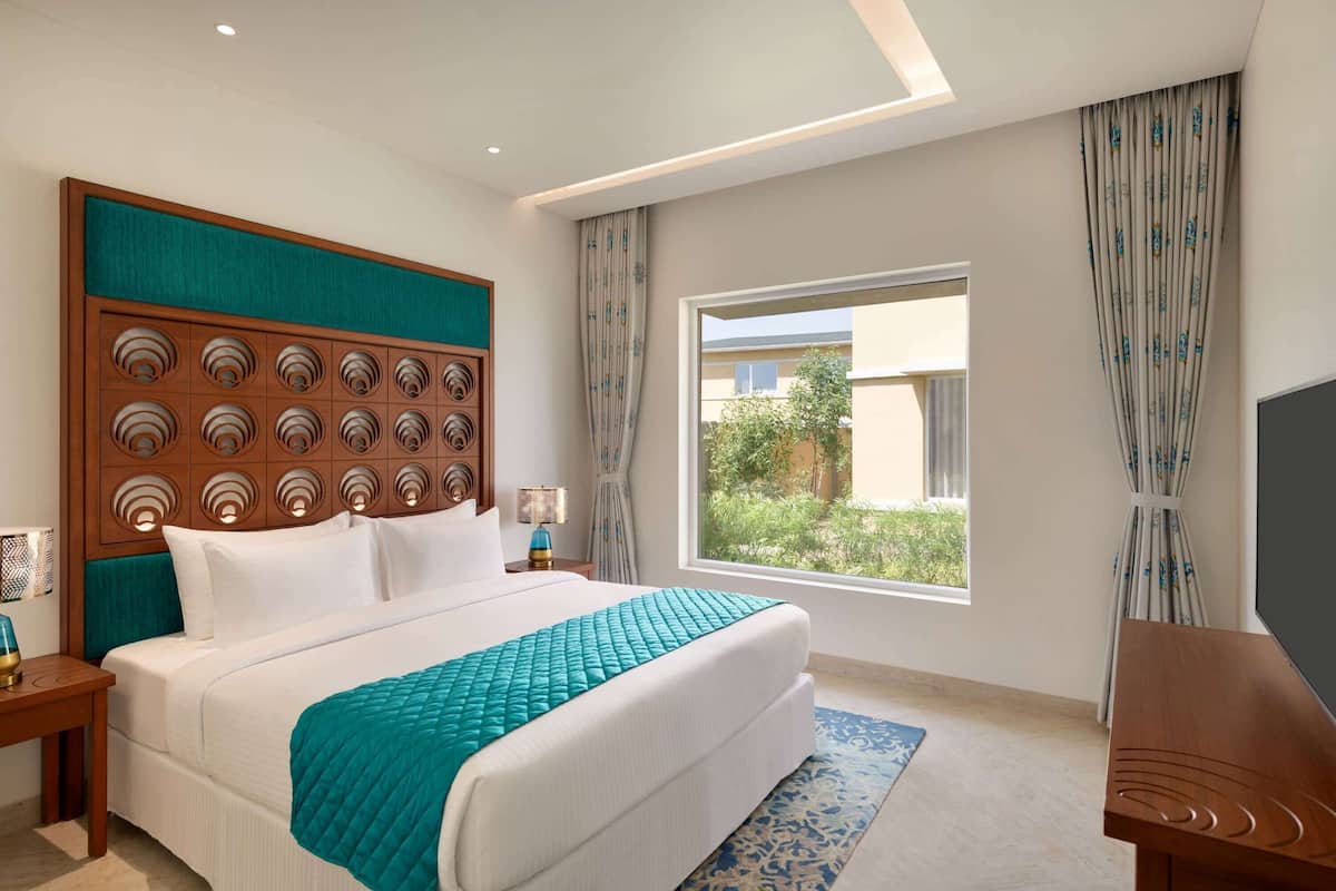 Habitación de hotel con una cama grande, cabecera de madera, televisor y ventana con cortinas.