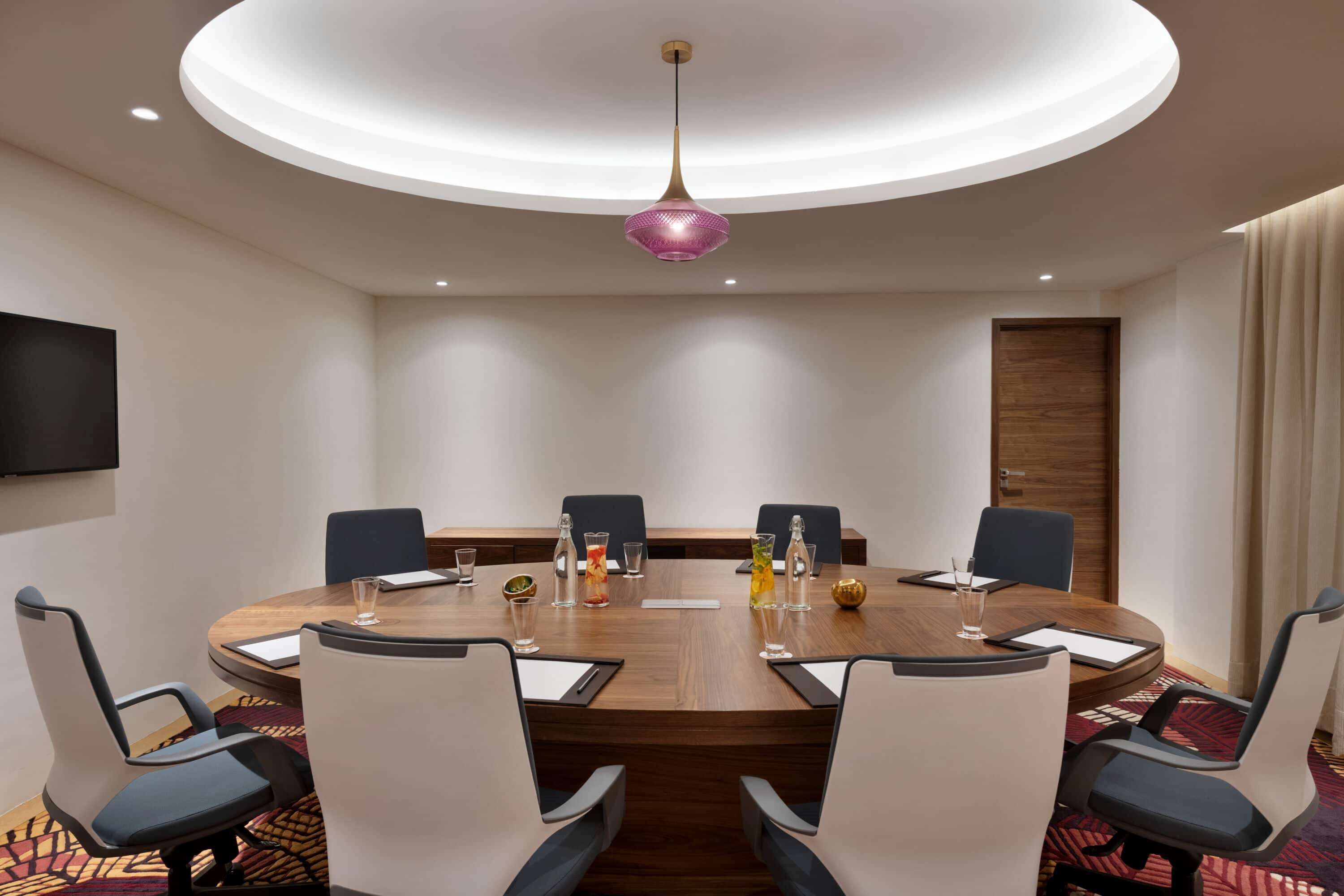 A conference room with a round table, six chairs, a modern pendant light, and a flat-screen TV on the wall.