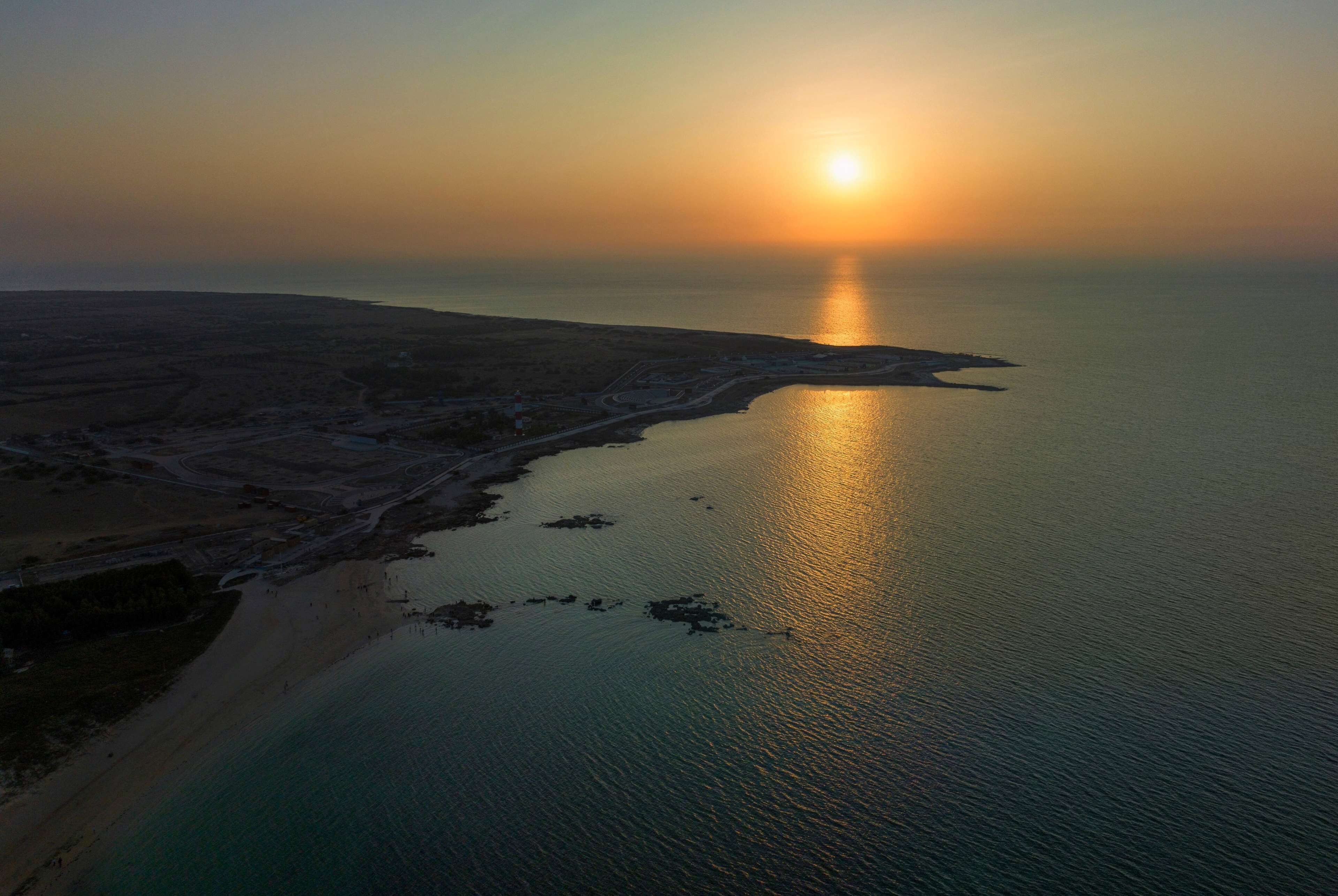 Vista aerea di una zona costiera con spiaggia e mare calmo che riflette il tramonto.