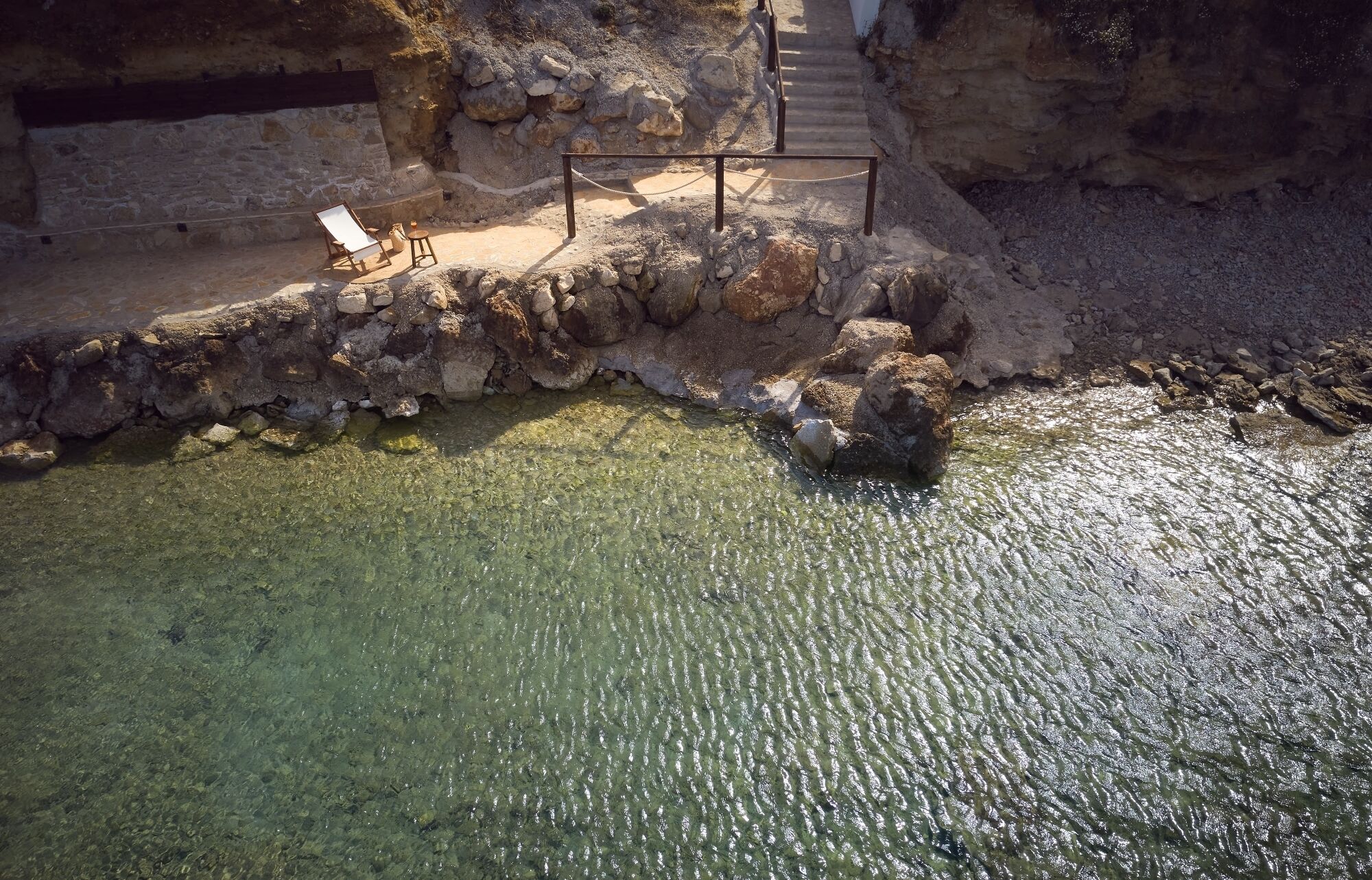 A stone pathway leading to a clear pool with a chair and table set up for relaxation.