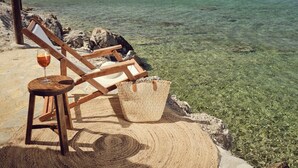 A wooden beach chair with a straw hat and a glass of drink on a stone surface by the sea. - Oceanis Luxury Suites (Zakynthos)