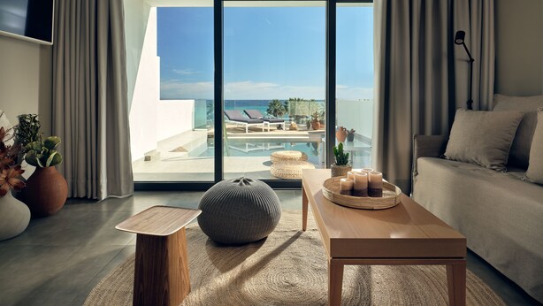 A modern living room with a large sliding glass door leading to a pool area, a comfortable sofa, a wooden coffee table, and decorative plants. - Oceanis Luxury Suites (Zakynthos)