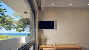 A room with a wooden desk, a chair, a flat-screen TV, and a view of the sea through a large window. - Oceanis Luxury Suites (Zakynthos)