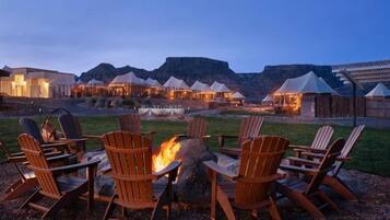 A fire pit with chairs, tents, and a building with lights in the background.