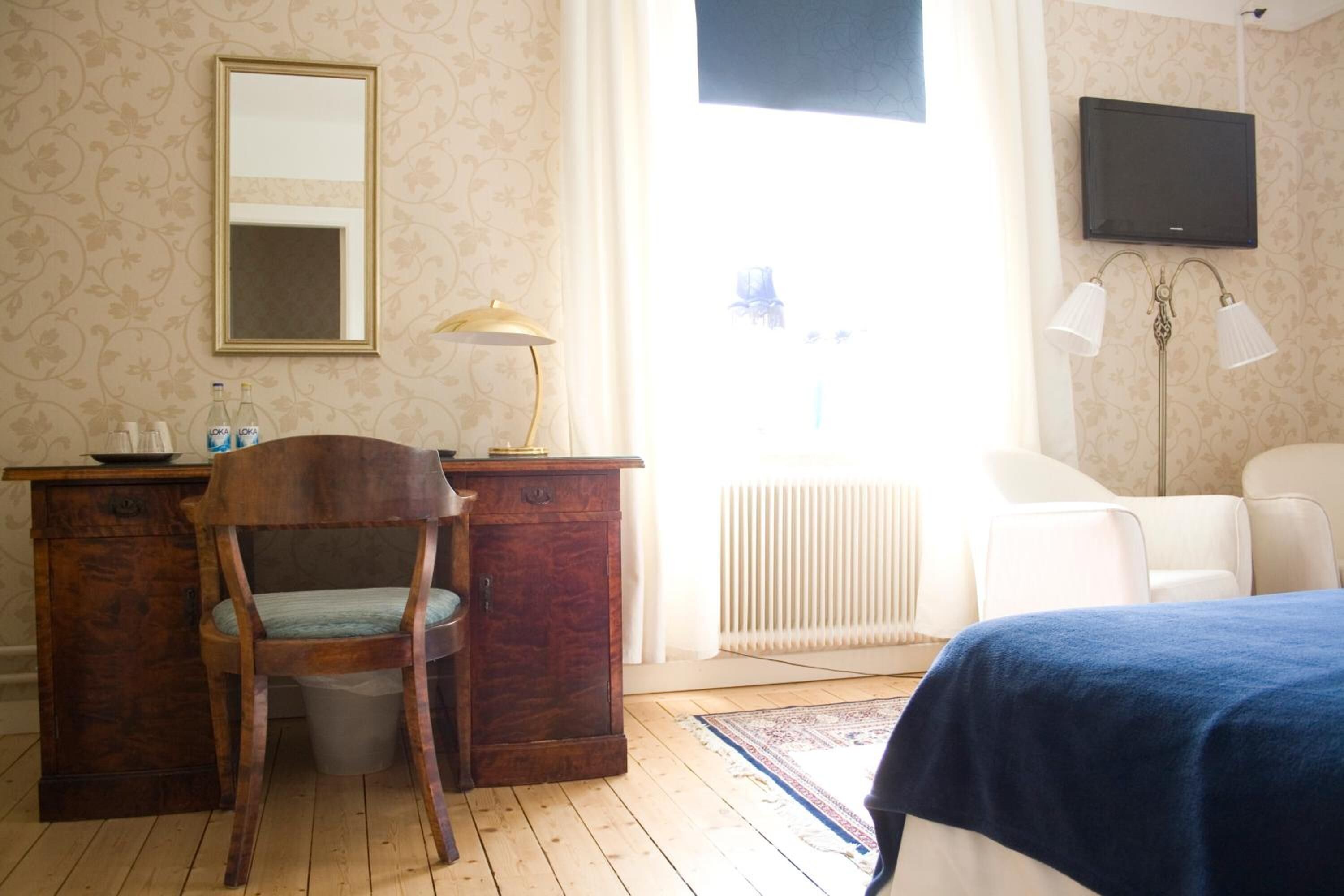 Een kamer met een houten bureau, een stoel, een bed met een blauwe plaid, een radiator, een televisie aan de muur en een raam met gordijnen.