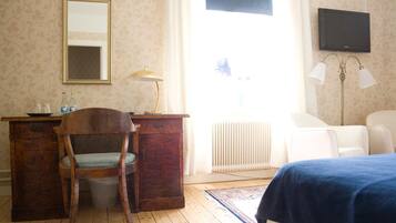 Een kamer met een houten bureau, een stoel, een bed met een blauwe plaid, een radiator, een televisie aan de muur en een raam met gordijnen.