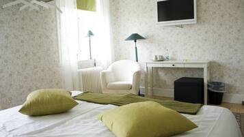 Habitación de hotel con una cama, dos almohadas verdes, una silla blanca, un escritorio con una computadora, un televisor y un radiador.
