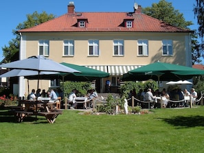 A building with a red-tiled roof and multiple windows. There are outdoor tables and chairs with green umbrellas, and people are seated and standing around. - Pensionat Prästgården (Töreboda)