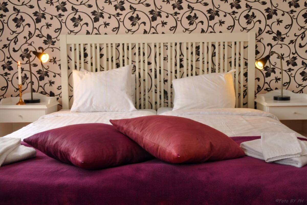 Ein ordentlich bezogenes Bett mit weißen Leinen und zwei roten Kissen, vor einer Wand mit Blumenmuster.