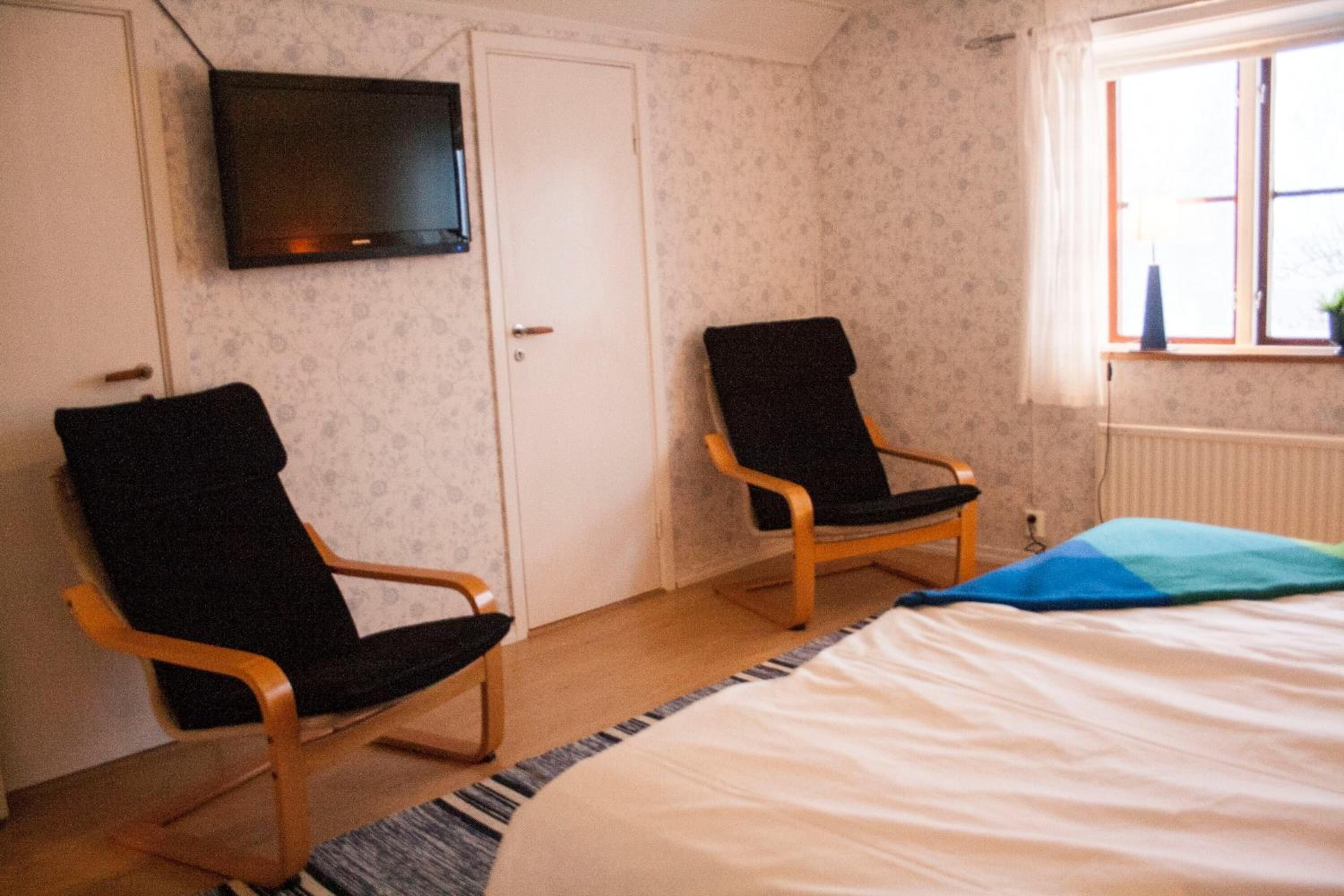 Un dormitorio con dos sillas de madera, una cama, un televisor en la pared y una ventana con vistas.
