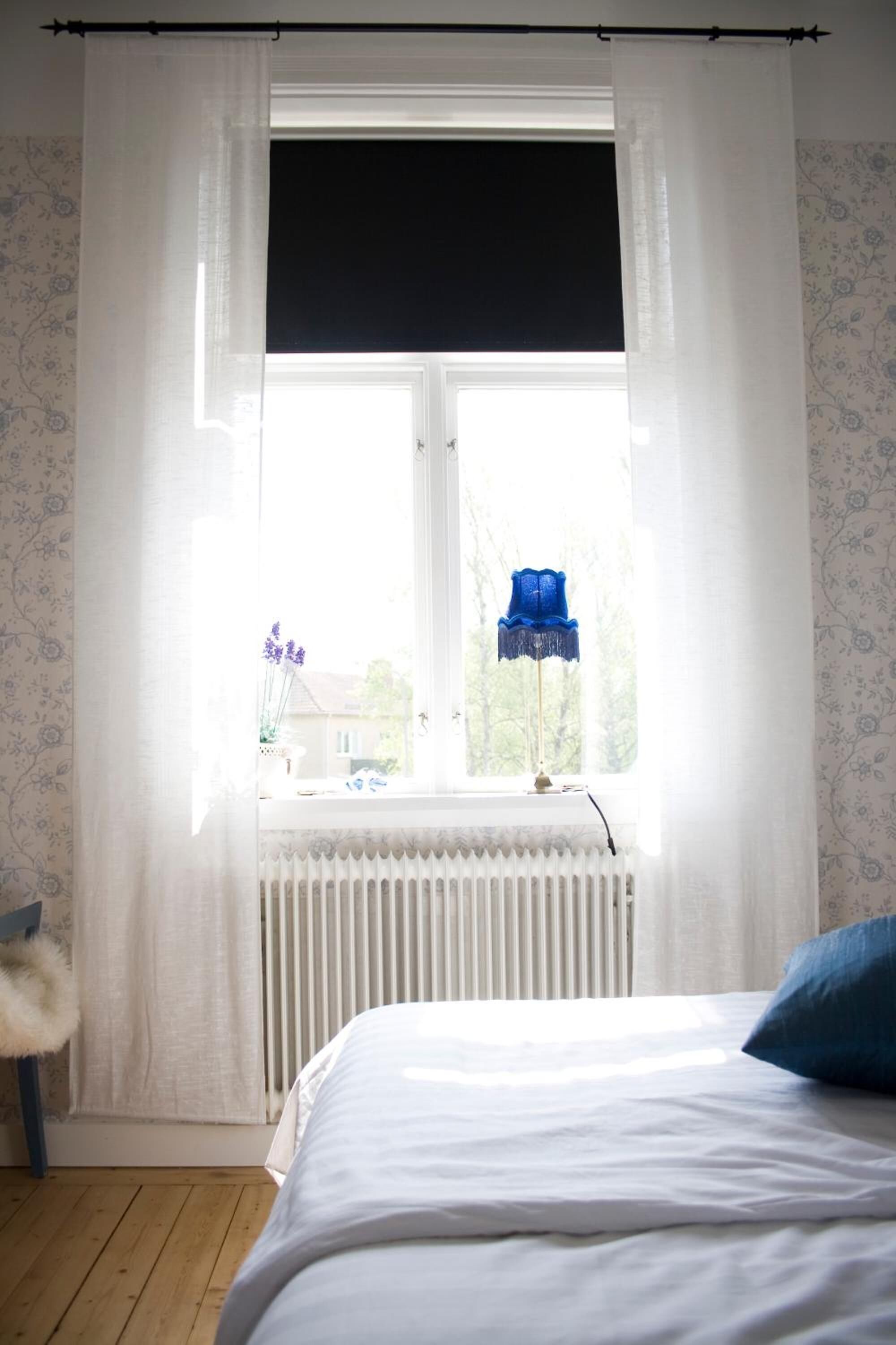 Een slaapkamer met een bed, een raam met dunne gordijnen, een radiator en een blauwe lamp op de vensterbank.