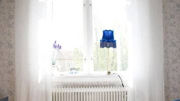 Een slaapkamer met een bed, een raam met dunne gordijnen, een radiator en een blauwe lamp op de vensterbank.