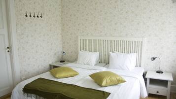 A neatly made bed with white bedding and two green pillows, a white bedside table with a lamp, and a wall with floral wallpaper.