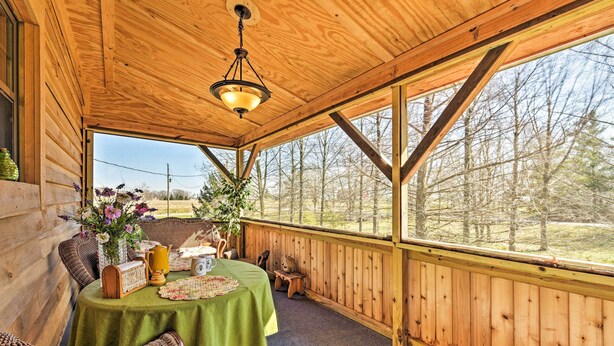 A wooden deck with a table set for dining, surrounded by wicker chairs, and a view of trees through large windows.