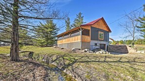 A cabin with a wooden deck, a stone foundation, and a red roof, surrounded by trees and a grassy area.