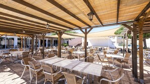 Un café en plein air avec des tables et des chaises en osier, installé sous une pergola en bois et doté d’un sol carrelé.