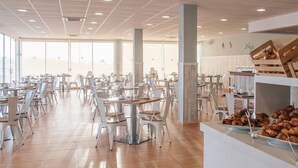 Un vaste espace dédié à la salle à manger, avec de nombreuses tables et chaises, un buffet proposant des pâtisseries et de grandes fenêtres laissant entrer la lumière naturelle.
