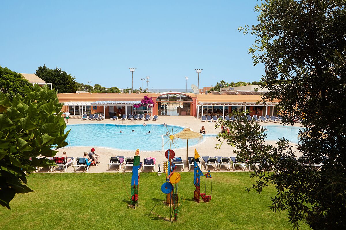 Ein Poolbereich mit mehreren Liegestühlen und Sonnenschirmen, umgeben von einem Gebäude mit einem überdachten Eingang.