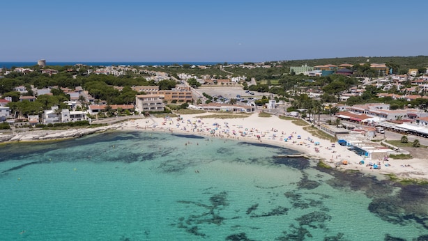 Vue aérienne d’une ville côtière avec une plage de sable, des eaux turquoise et limpides, et un mélange de bâtiments résidentiels et commerciaux.