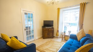 A living room with two sofas, a television on a cabinet, a large window with curtains, and a door with glass panels. - Sandgate 2-bed Apartment in Ayr Central Location (Ayr)