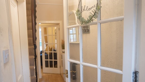 A hallway with a wooden floor, a black doormat, a white door with frosted glass panels, and a chandelier. - Sandgate 2-bed Apartment in Ayr Central Location (Ayr)