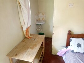 Una habitación pequeña y sencilla con una mesa de madera, una cama con un solo almohadón, una cesta verde y un lavamanos básico.