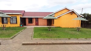 A building with a yellow exterior and a red roof, featuring a paved entrance and two outdoor tables with umbrellas. - Eastward Garden Hotel (Nyahururu)