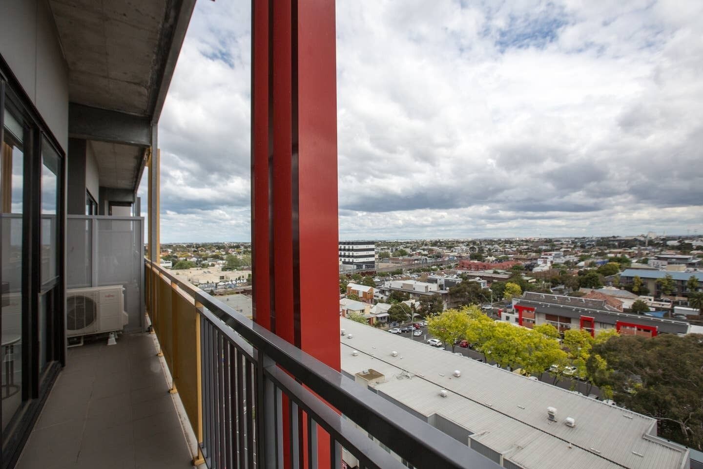A balcony with a clear view of a residential area, a red structural column, and a building with multiple stories.