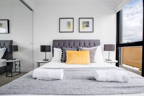 Una habitación de hotel moderna con una cama grande, dos mesitas de noche, un cabecero tapizado en gris y dos cuadros enmarcados en la pared.