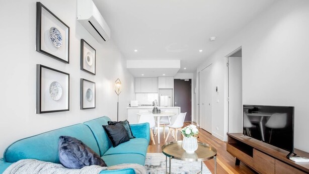 A modern living room with a blue sofa, a coffee table, and a TV. There is a kitchen in the background with white cabinets and a dining area. - Elegant 2br@footscray Station (Footscray)