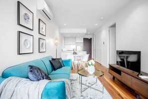 Una sala moderna con un sofá azul, una mesa de centro y un televisor. Se ve una cocina al fondo con gabinetes blancos y un comedor.