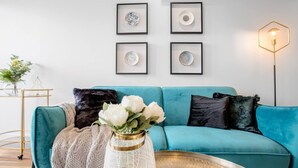 A teal sofa with white and black cushions, a white vase with flowers, and a floor lamp. - Elegant 2br@footscray Station (Footscray)