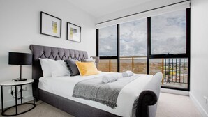 A modern bedroom with a large bed, a side table, a lamp, and a view of the city through large windows. - Elegant 2br@footscray Station (Footscray)