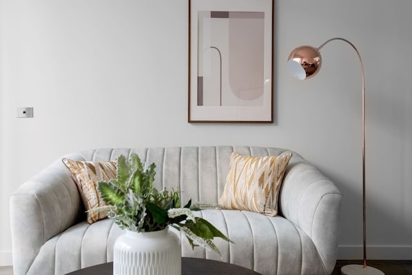 A living room with a grey sofa, two patterned pillows, a white vase with green plants, and a floor lamp.