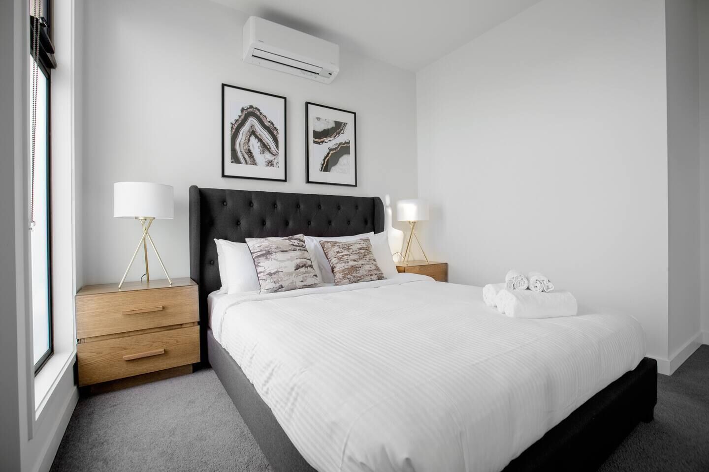 A modern bedroom with a large bed, two bedside tables with lamps, a dark headboard, and two framed abstract paintings on the wall.