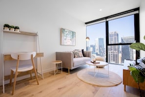 Un salon moderne comprenant un canapé, une table basse ronde, une petite table d’appoint et un fauteuil. Une grande fenêtre offre une vue sur la ville.