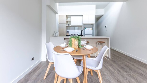 A modern kitchen with white cabinets, a central island, and a dining area with a round table and white chairs. - Elegant Stylish Living@mooneeponds Wifi/parking (Moonee Ponds)