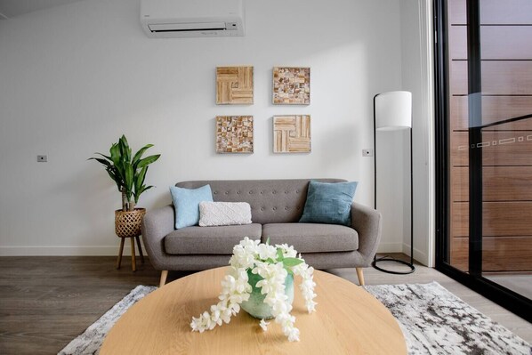 Un salon moderne avec un canapé gris, des coussins décoratifs, des œuvres d’art murales, une plante en pot et une table basse en bois agrémentée de fleurs.
