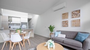 A modern living space with a grey sofa, white chairs, a round wooden coffee table, and a kitchen area with white cabinets and a built-in oven. - Elegant Stylish Living@mooneeponds Wifi/parking (Moonee Ponds)