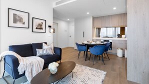 Un soggiorno moderno con un divano blu scuro, un tavolino da caffè e un'area pranzo con sedie blu. Alle pareti si notano quadri incorniciati e sullo sfondo si intravede una cucina.