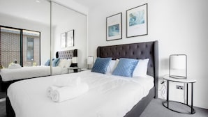 A modern hotel room with a large bed, a smaller bed, a headboard, a bedside table with a lamp, and artwork on the wall.