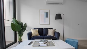 A modern living room with a dark blue sofa, a white coffee table, a large green plant, and a framed abstract painting on the wall.