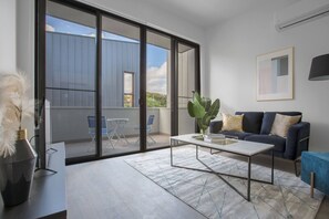 A modern living room with a sofa, coffee table, and a view of a blue building through large glass doors. - Trendy Livingstyle 2br@moonee Ponds Wifi+parking (Moonee Ponds)