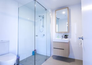 A modern bathroom with a glass-enclosed shower, a white toilet, a wooden vanity with a mirror, and a small potted plant. - Stylist 2 Bedrooms Apartment With Pool/gym/parking (South Brisbane)