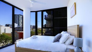 Ein modernes Schlafzimmer mit einem großen Bett, einem Nachttisch, einer Lampe und Blick auf die Stadt durch raumhohe Fenster.